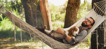 Mies makaa riippumatossa silmät kiinni koira sylissään. Taustalla puidenrunkoja, joiden välistä paistaa päivä.