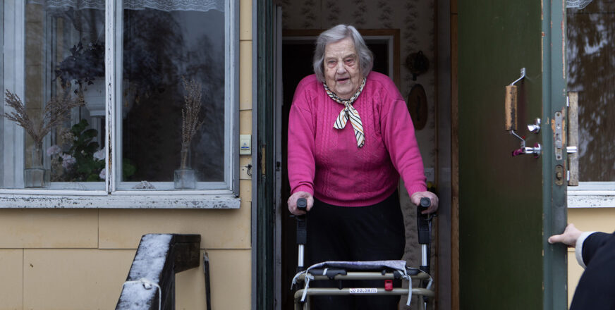 Hilkka ulko-ovellaan rollaattorin kanssa iloisena vastaanottaa vieraita.